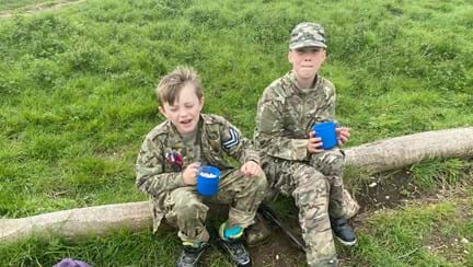 Two boys sat on a log wearing camouflage clothing 