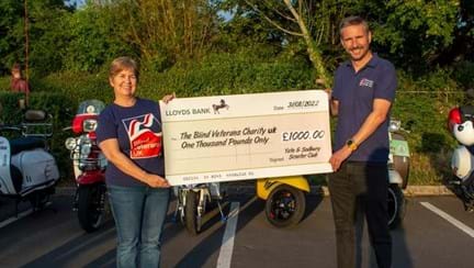 Sally and Colin in their Blind Veterans UK T-shirts holding a giant cheque from the scooter club