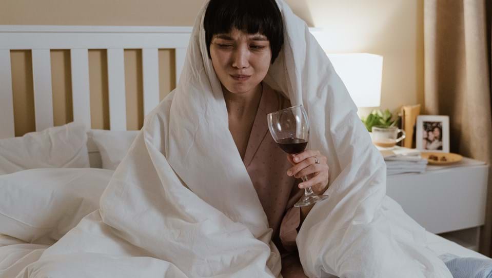 A woman with a scrunched-up face, sitting in bed, wrapped in a duvet, looking at her laptop, while drinking wine and eating chocolates