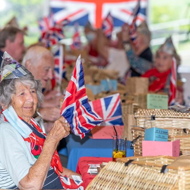 Several blind veterans holding up British flags, and wearing party hats, celebrating together at a long table with drinks and food