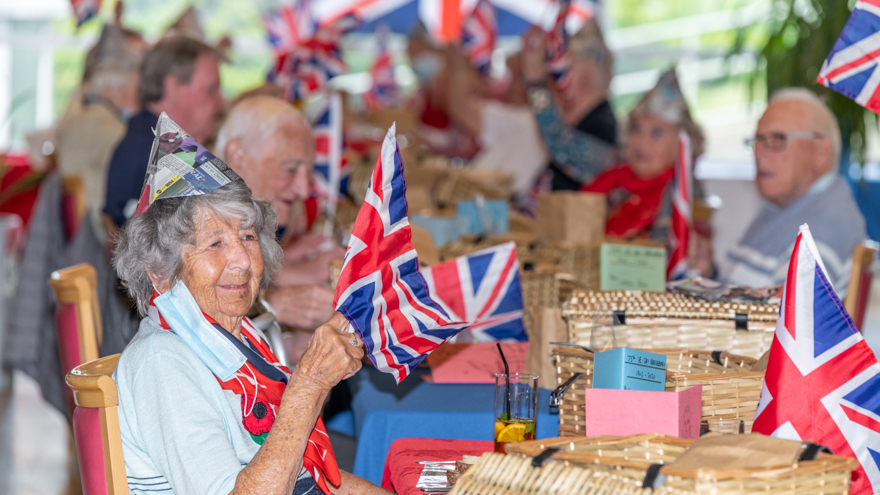 Several blind veterans holding up British flags, and wearing party hats, celebrating together at a long table with drinks and food