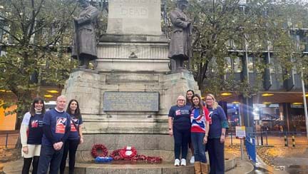 Seven members of Thea staff wearing Blind Veterans UK t shirts stood around the war memorial with poppy wreaths laid at its base