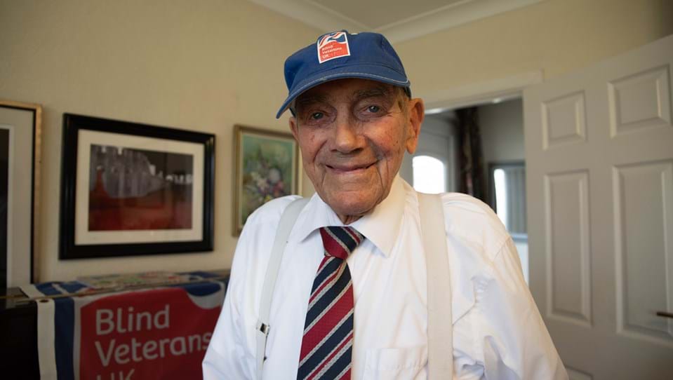 Blind veteran Eddie looking into the camera smiling while wearing a Blind Veterans UK cap and tie