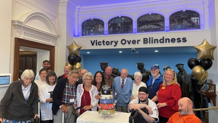 A group of Llandudno staff and visiting veterans stood in front of the Llandudno Centre's Victory over Blindness statue. They are surrounding the five tiered celebratory cake.