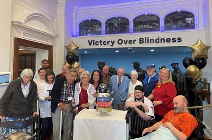A group of Llandudno staff and visiting veterans stood in front of the Llandudno Centre's Victory over Blindness statue. They are surrounding the five tiered celebratory cake.