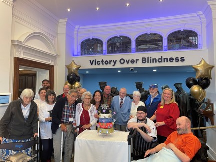 A group of Llandudno staff and visiting veterans stood in front of the Llandudno Centre's Victory over Blindness statue. They are surrounding the five tiered celebratory cake.
