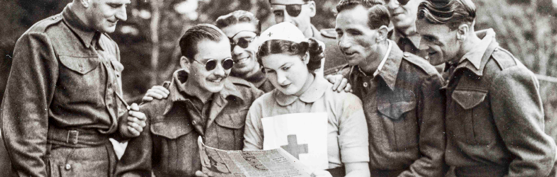 A nurse standing while reading a newspaper, as a group of blind veterans are gathered round her 