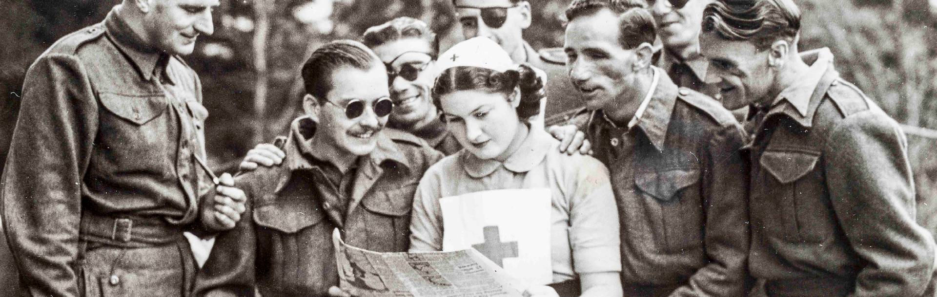 A nurse standing while reading a newspaper, as a group of blind veterans are gathered round her