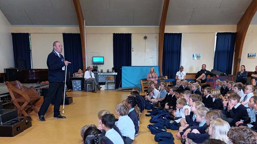 Blind veteran Billy standing in front of a group of pupils and teachers giving a talk in a school hall