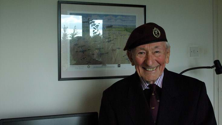 Blind veteran Jim smiling as he stands in front of a framed piece of artwork showing the Horsa Gilders landing at Arnhem
