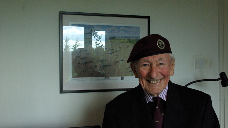 Blind veteran Jim smiling as he stands in front of a framed piece of artwork showing the Horsa Gilders landing at Arnhem