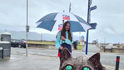 A photo of Emmie, standing next to the Llandudno's Cheshire Cat statue, holding a Blind Veterans UK umbrella