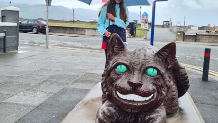 A photo of Emmie, standing next to the Llandudno's Cheshire Cat statue, holding a Blind Veterans UK umbrella