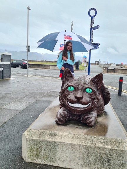 A photo of Emmie, standing next to the Llandudno's Cheshire Cat statue, holding a Blind Veterans UK umbrella
