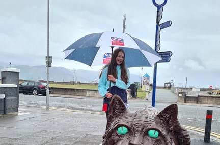 A photo of Emmie, standing next to the Llandudno's Cheshire Cat statue, holding a Blind Veterans UK umbrella