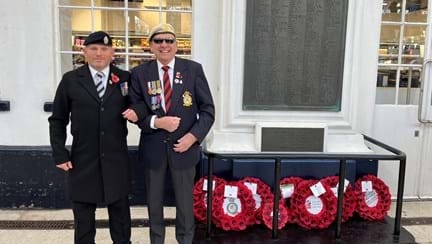 Tony and Danny arm in arm and smiling stood to the side of the war memorial at Brighton Train Station which has eight poppy wreaths laid at its base