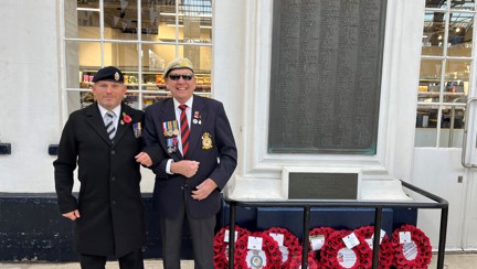 Tony and Danny arm in arm and smiling stood to the side of the war memorial at Brighton Train Station which has eight poppy wreaths laid at its base