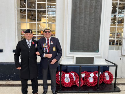 Tony and Danny arm in arm and smiling stood to the side of the war memorial at Brighton Train Station which has eight poppy wreaths laid at its base