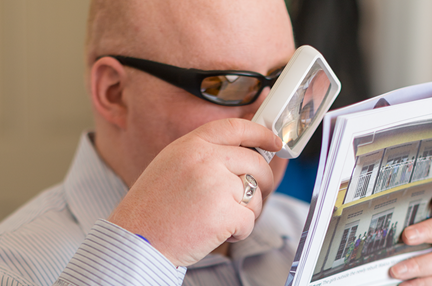 A blind veteran wearing dark glasses, holding up a hand magnifier to his eye to read a magazine