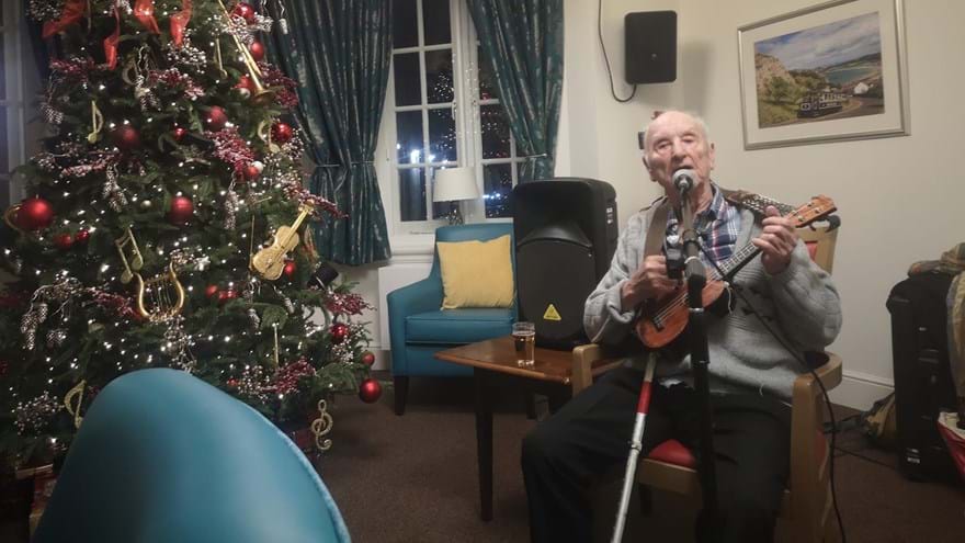 A man sitting next to a large Christmas tree playing the ukelele and singing