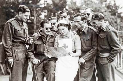 A nurse standing while reading a newspaper, as a group of blind veterans are gathered round her
