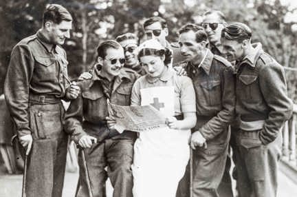 A nurse standing while reading a newspaper, as a group of blind veterans are gathered round her