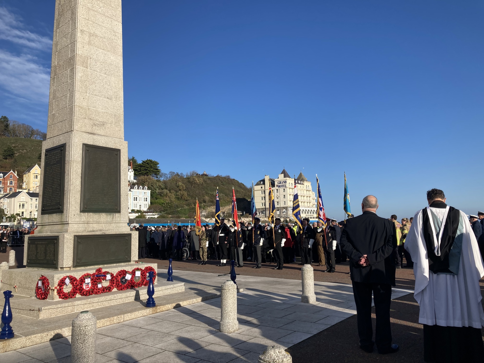 Llandudno town war memorial with poppy wreaths at its base. Standard bearers and veterans can be seen looking towards the memorial in the background.