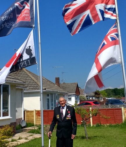 Blind veteran Eddie standing between two flag poles in his garden, displaying a Blind Veterans UK, Union Jack and Lest We Forget flag 