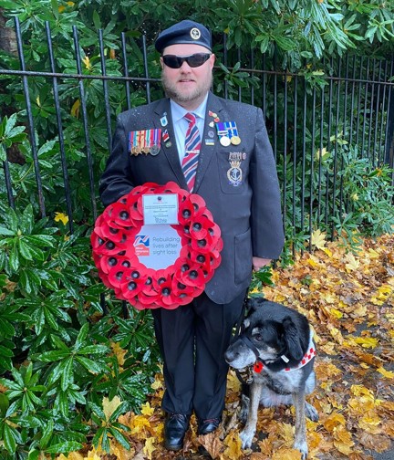 Colin in his suit with military medals holding a poppy wreath which has a label attached as it has been gifted by Thea. Colin's guide dog Diamond is sat beside him