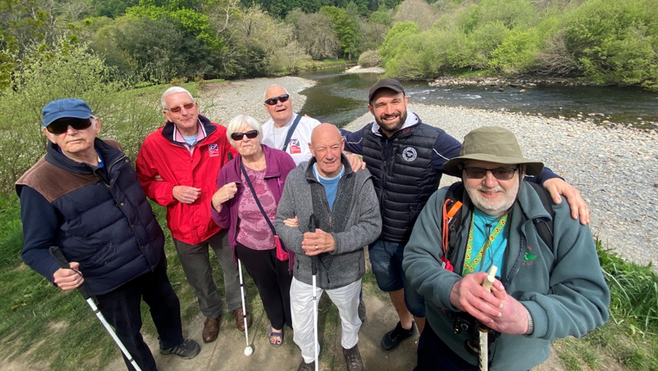 Blind veterans, smiling enjoying a group walk at the Llandudno centre. Some have canes. They are by a river with trees in the background