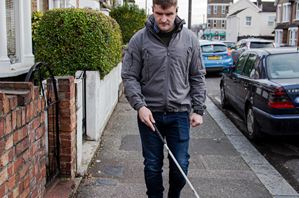 Blind veteran Rob walking along a pavement, using a long white cane to guide him