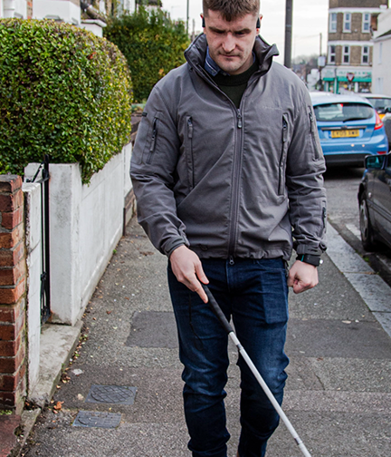 Blind veteran Rob walking along a pavement, using a long white cane to guide him