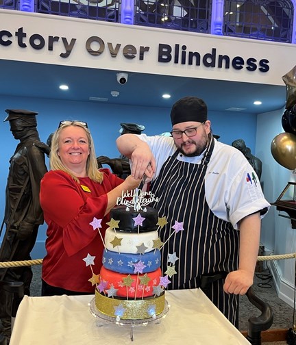 Kathy and Joe stood in front of the Victory over Blindness sculpture at the Llandudno Centre. They are ready to cut the five tier cake covered with stars and a cake topper that reads "Well done Llandudno"