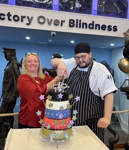 Kathy and Joe stood in front of the Victory over Blindness sculpture at the Llandudno Centre. They are ready to cut the five tier cake covered with stars and a cake topper that reads "Well done Llandudno"