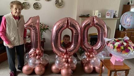 Blind veteran Margaret stands beside huge pink balloons that read '100'
