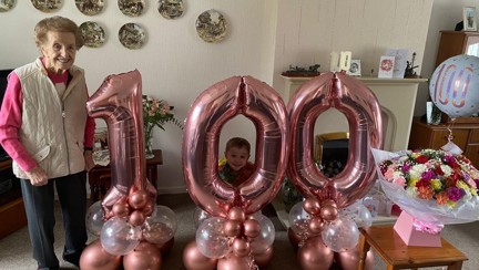 Blind veteran Margaret stands beside huge pink balloons that read '100'