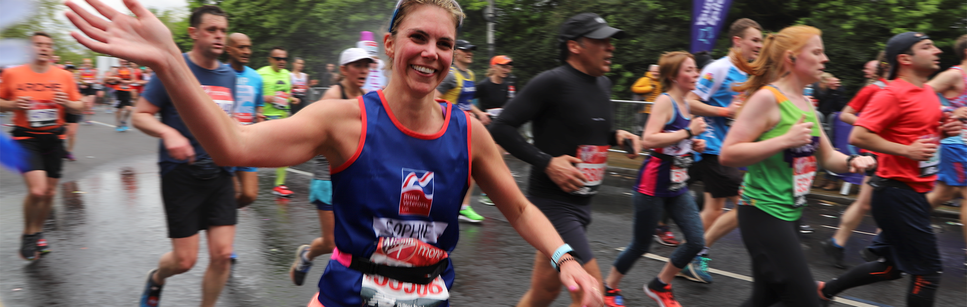 Charity supporter, Sophie Delaney, wearing a Blind Veterans UK vest, running alongside a crowd of marathon runners, smiling and waving as she passes the camera.
