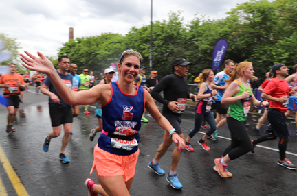 Charity supporter, Sophie Delaney, wearing a Blind Veterans UK vest, running alongside a crowd of marathon runners, smiling and waving as she passes the camera.