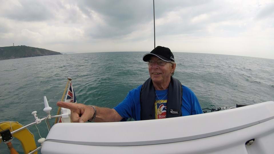 Blind veteran Rod on a sailing boat with the sea behind him, smiling as he and points ahead