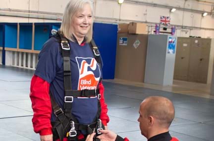 Christine wearing a Blind Veterans UK T-shirt over her red overalls with a man in red overalls knelt down tightening up her harness