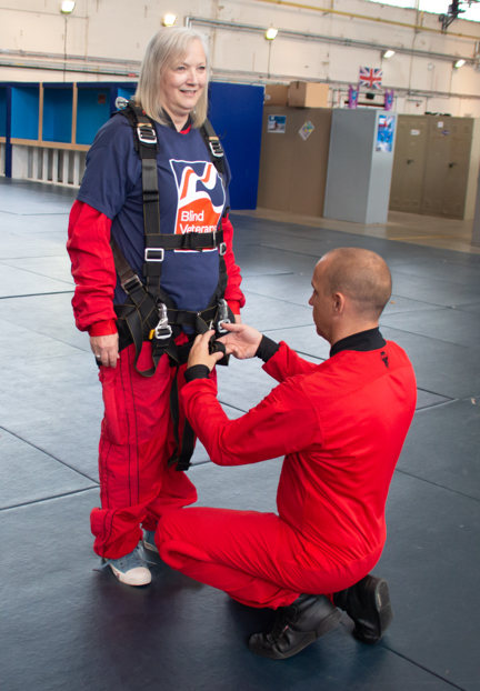 Christine wearing a Blind Veterans UK T-shirt over her red overalls with a man in red overalls knelt down tightening up her harness