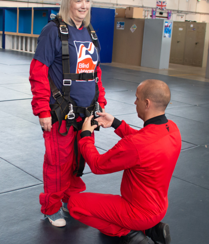 Christine wearing a Blind Veterans UK T-shirt over her red overalls with a man in red overalls knelt down tightening up her harness