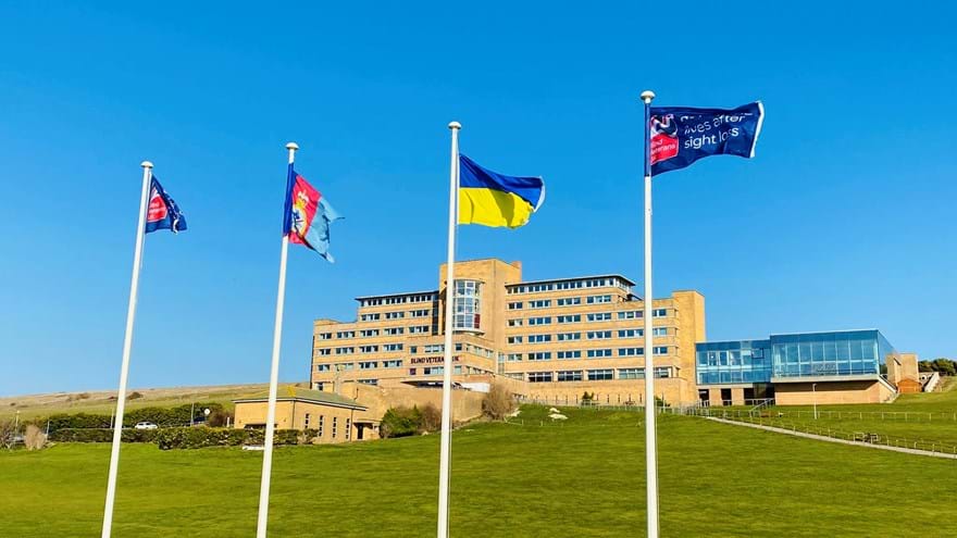 Four tall flagpoles with flags flying, at the bottom of a grassy hill, with our Centre of Wellbeing in Brighton in the background