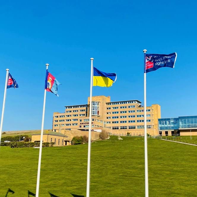 Four tall flagpoles with flags flying, at the bottom of a grassy hill, with our Centre of Wellbeing in Brighton in the background