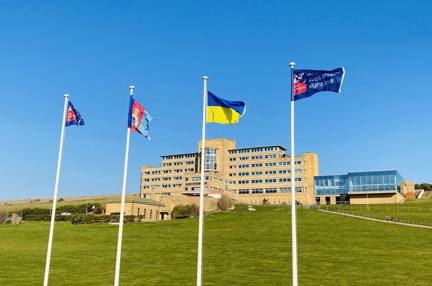 Four tall flagpoles with flags flying, at the bottom of a grassy hill, with our Centre of Wellbeing in Brighton in the background