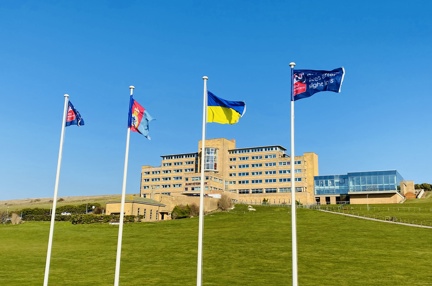 Four tall flagpoles with flags flying, at the bottom of a grassy hill, with our Centre of Wellbeing in Brighton in the background