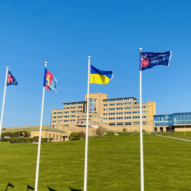 Four tall flagpoles with flags flying, at the bottom of a grassy hill, with our Centre of Wellbeing in Brighton in the background