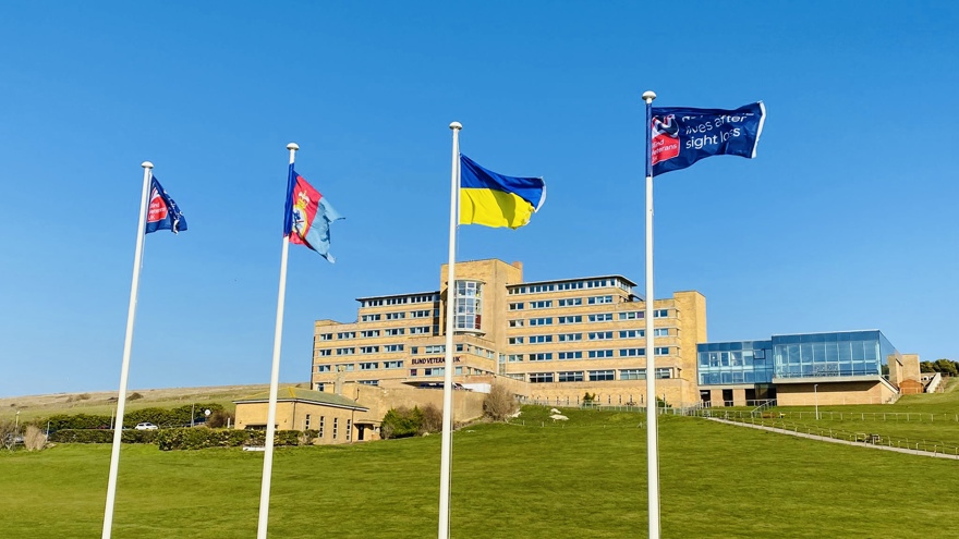 Four tall flagpoles with flags flying, at the bottom of a grassy hill, with our Centre of Wellbeing in Brighton in the background