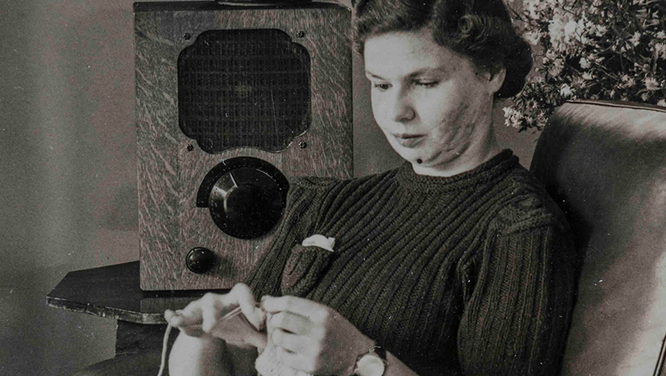 An archive photograph of a young blind veteran Barbara sitting on an armchair and sewing