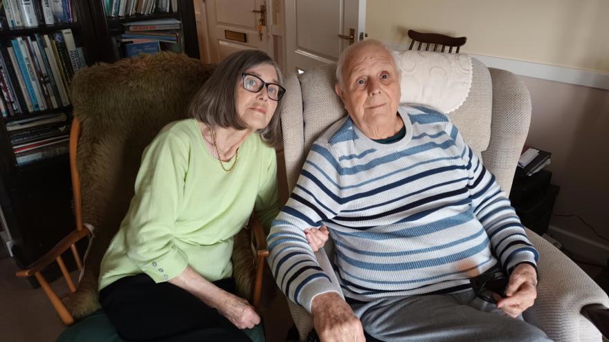 A photo of blind veteran Dudley, right, and his wife Yvonne, left sitting on chairs while linking arms